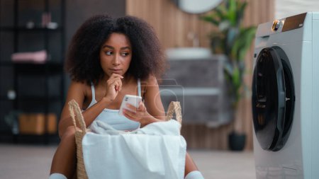 Photo for African American female woman girl pensive thinking ponder idea thoughtful think solution hesitate decide using mobile phone online shopping smartphone at floor with laundry basket washing machine - Royalty Free Image