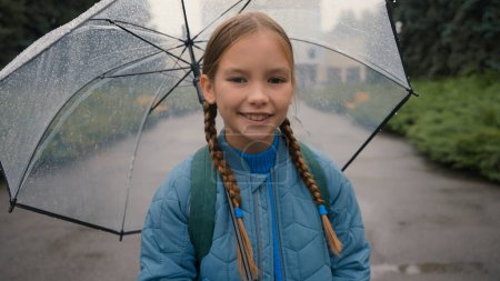 Téléchargez les photos : Petite fille européenne regardant la caméra souriant tenant parapluie pluie météo parc marcher à l'extérieur fille enfant apprenant enfant élève dent sourire joie plaisir positif femelle humide saison gouttelettes climat - en image libre de droit