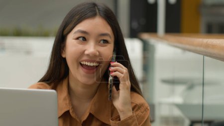 Foto de Mujer asiática sonriente empresaria que trabaja en línea con el ordenador portátil en la oficina chica japonesa china estudiante freelancer hablar teléfono móvil riendo emocionado respuesta femenina teléfono inteligente llamada conversación despreocupada - Imagen libre de derechos