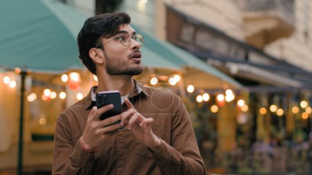 Photo for Nervous lonely Indian Arabian ethnic male businessman guy looking around feeling lost. Man holding smartphone use mobile electronic map geolocation app trying find location outside city evening street - Royalty Free Image
