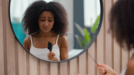 Frustrated African American woman suffer problem trouble curls looking at mirror at bathroom beauty routine prepare biracial girl female dry hair trying to brush combing curly tangled hairdo with comb