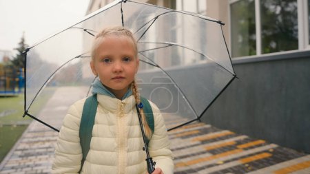 Téléchargez les photos : Écolière petite fille va tenant parapluie pluie temps humide nuageux après les leçons élémentaire promenade enfant fille fille enfant femelle élève camarade de classe week-end éducatif étude regardant caméra - en image libre de droit