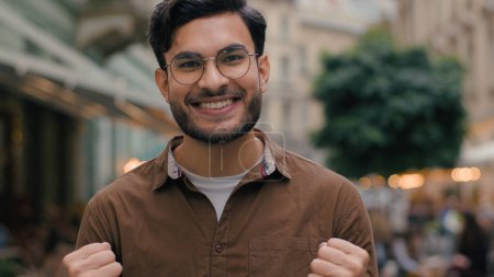 Photo for Excited Indian Arabian ethnic male man 20s guy bearded businessman happy student celebrating achievement victory clenching fists gesture good news winning lottery looking at camera outside city street - Royalty Free Image