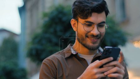 Photo for Close up successful Indian Arabian ethnic man male student gen z guy businessman happy smartphone user chatting scrolling mobile phone device online application chat walking night city street outdoors - Royalty Free Image