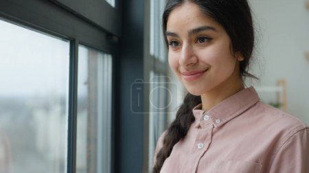 Photo for Arabian woman female lady dreaming homeowner businesswoman calm girl walking near window glass panorama thoughtful walk at home looking outside staring look out city view waiting think dream memories - Royalty Free Image