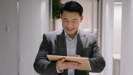 Téléchargez les photos : Heureux Asiatique d'âge moyen ethnique homme chinois coréen homme d'affaires souriant employeur d'entreprise utiliser tablette numérique dans le couloir de bureau lobby entrepreneur masculin navigation sur Internet médias sociaux sur appareil portable - en image libre de droit