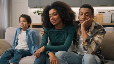 Sad African American family desperate parents hug unhappy mother woman female dissatisfied dad man male sit couch ignore misbehaves little child loud shouting boy active kid son jumping sofa room home