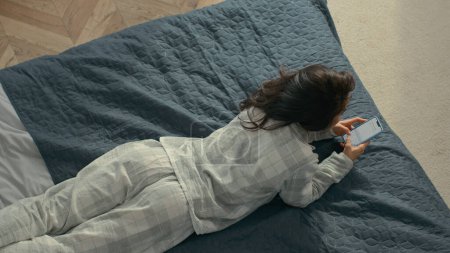 Téléchargez les photos : Top vue jeune femme brune joyeuse en pyjama couché sur le lit confortable maison à l'intérieur chambre à coucher femelle rouler sur le chat textos smartphone téléphone mobile communiquer sourire week-end joie - en image libre de droit