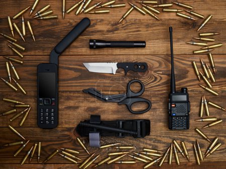 Close-up on a wooden background. Satellite phone with a large antenna, radio station, fixed-blade knife, flashlight, atraumatic scissors, and hemostatic tourniquet. Frame of cartridges.