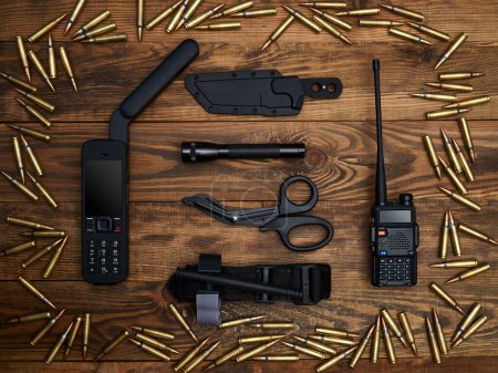 Close-up on a wooden background. Satellite phone with a large antenna, radio station, tactical knife in a sheath, flashlight, atraumatic scissors, and hemostatic tourniquet. Frame of cartridges.