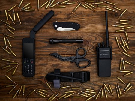 Close-up on a wooden background. Satellite phone with large antenna, radio station, tactical knife, pocket knife, flashlight, atraumatic scissors, hemostatic tourniquet. Frame of cartridges.
