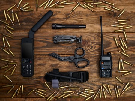 Close-up on a wooden background. Satellite phone with a large antenna, radio station, multi-tool, flashlight, atraumatic scissors, and hemostatic tourniquet. Frame of cartridges.