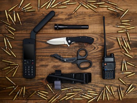 Close-up on a wooden background. Satellite phone with a large antenna, radio station, fixed-blade knife, flashlight, atraumatic scissors, and hemostatic tourniquet. Frame of cartridges.