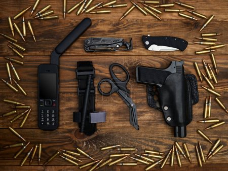 Close-up on a wooden background. Satellite phone with a large antenna, multi-tool, handgun in a sheath, pocket knife, atraumatic scissors, and hemostatic tourniquet. Frame of cartridges.