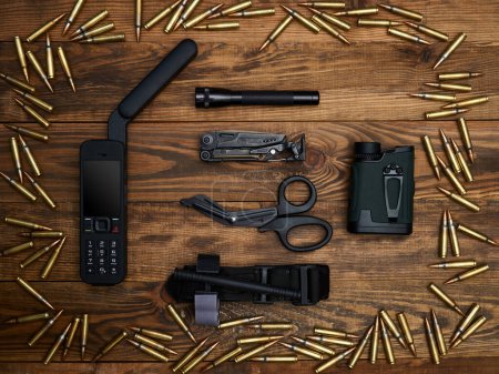 Close-up on a wooden background. Satellite phone with a large antenna, multi-tool, flashlight, atraumatic scissors, laser rangefinder, and hemostatic tourniquet. Frame of cartridges.