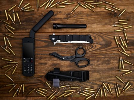 Close-up on a wooden background. Satellite phone with a large antenna, tactical knife, flashlight, atraumatic scissors, and hemostatic tourniquet. Frame of cartridges.