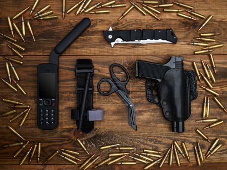Close-up on a wooden background. Satellite phone with a large antenna, tactical knife, handgun, atraumatic scissors, and hemostatic tourniquet surrounded by a frame of cartridges
