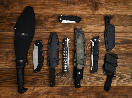 Close-up of nine different knives with closed blades or in sheaths. All laid out on the brown wooden background. Fixed-blade, pocket, and tactical knives. Black handles.