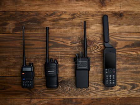 Closeup of an assortment of three black radio stations of different sizes and a satellite phone with a large antenna, laid out on a brown wooden background