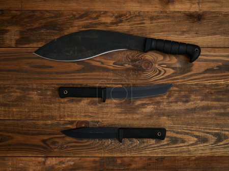Close-up of three different fixed-blade knives with black blades and black handles. All laid out on a brown wooden background.