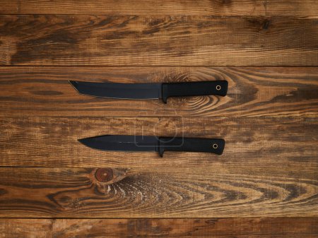 Close-up of two fixed-blade knives with black blades and black handles laid out on a brown wooden background.