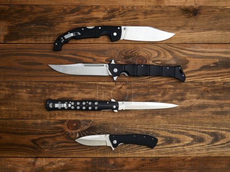 Close-up of four folding knives laid out on the brown wooden background. Silver blades and black handles.