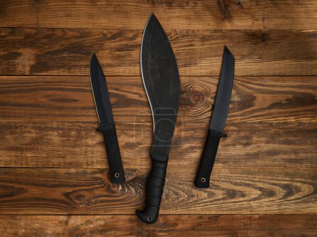 Gros plan de trois couteaux à lame fixe différents avec des lames noires et des poignées noires. Tous disposés sur un fond en bois brun.