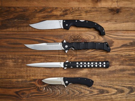 Close-up of four folding knives laid out on the brown wooden background. Silver blades and black handles.