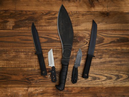 Gros plan de cinq différents couteaux à lame fixe disposés sur le fond brun en bois. Lames noires et argentées, poignées noires.