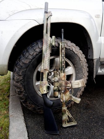 A shotgun and a rifle gun standing against the wheel of a silver SUV