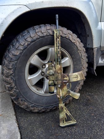 A camouflage-colored rifle gun standing against a mud-covered wheel of a silver SUV