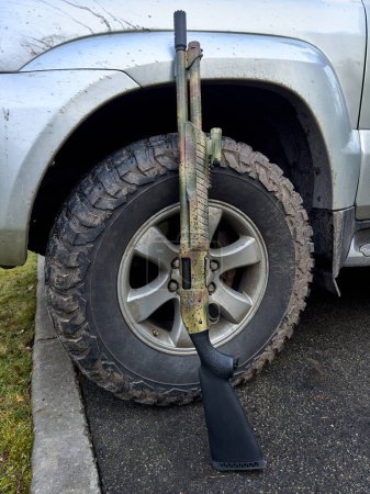 A camouflage-colored shotgun gun standing against a mud-covered wheel of a silver SUV