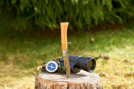 Photo for A rustic knife with a wooden handle, a vintage monocular, and a handheld compass rest on a wooden stump against a blurred background of lush green grass and towering pines. - Royalty Free Image