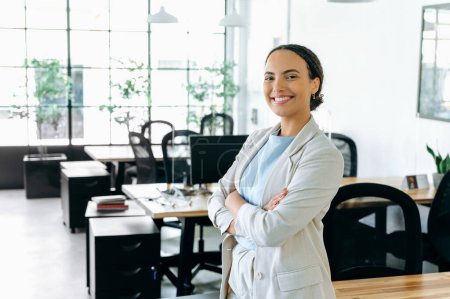 Photo for Confident successful brazilian or hispanic business woman, company employee, manager, in a jacket, standing with her arms crossed against the backdrop of the office, looking at the camera, smiling - Royalty Free Image