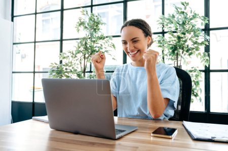 Photo for Amazed happy brazilian or hispanic business woman, sit at a desk with laptop in modern office, rejoicing in success, big profit, celebrate deal, job promotion, gesturing fists, looks at camera, smiles - Royalty Free Image