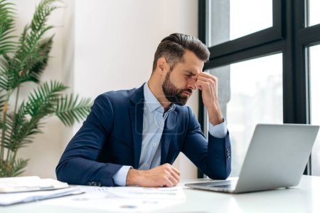 Frustrated overworked caucasian businessman, entrepreneur, product manager, programmer, sits at a desk in the office, massaging the bridge of his nose with his eyes closed, exhausted from long work