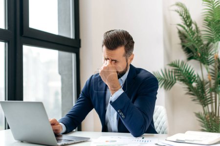 Overworking. Frustrated tired caucasian entrepreneur, product manager, programmer, sit at a desk in the office, massaging the bridge of his nose with his eyes closed, feeling exhausted from long work