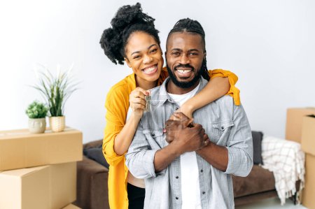 New housing, relocation. Happy joyful african american family, black couple hugging while standing in living room, woman showing keys of their new property, they looking at camera, smiling