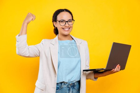 Photo for Successful mixed race business woman, company employee, with glasses, holds an open laptop, stand on isolated yellow background, looks at camera, smiles, demonstrates his strength with a hand gesture - Royalty Free Image