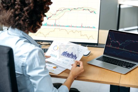 Photo for Back view of successful african american female stock investor, broker, financial expert, sit at work desk, uses computer and charts, analyzes risks and prospects, rise or fall of cryptocurrency - Royalty Free Image