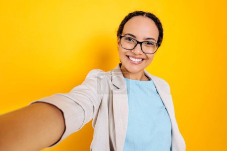 Gorgeous happy hispanic or brazilian curly haired woman with glasses, takes a selfie photo on her smartphone, looks at camera, smiles, stand on isolated yellow background