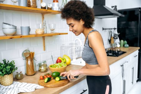 Photo for Happy african american young curly woman in a sportswear, standing at home in kitchen cutting cucumber while preparing fresh salad for healthy eating, smiling. Healthy food preparation concept - Royalty Free Image
