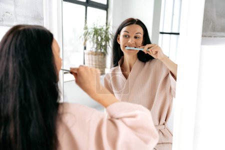 Schöne kaukasische Frau im Bademantel, die sich nach dem Schlafen vor einem Spiegel im Badezimmer die Zähne putzt und auf Mundhygiene achtet, um Karies zu vermeiden. Zahnpflege, Morgenroutine