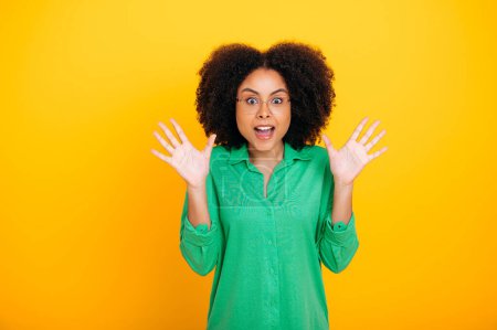 Amazed enthusiastic curly woman of brazilian or african american ethnicity, looks in surprise, joyfully at the camera, gesticulates with her palms with open mouth, stands on isolated yellow background