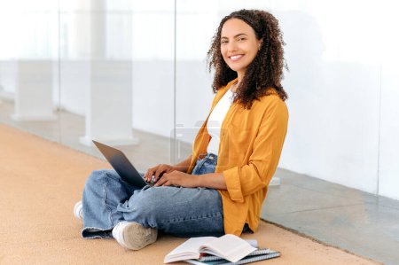 Using laptop. Education concept, female student. Beautiful stylish curly haired mixed race girl, sit near the university campus, using laptop, working on a project, looking at camera, smile friendly