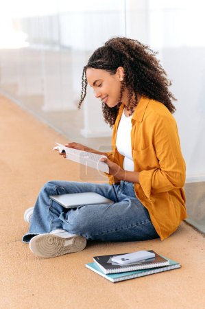 Photo verticale d'une belle étudiante brésilienne ou hispanique aux cheveux bouclés et positifs, assise près du campus universitaire, lisant un livre, se préparant à l'examen, faisant ses devoirs, s'inspirant, souriant