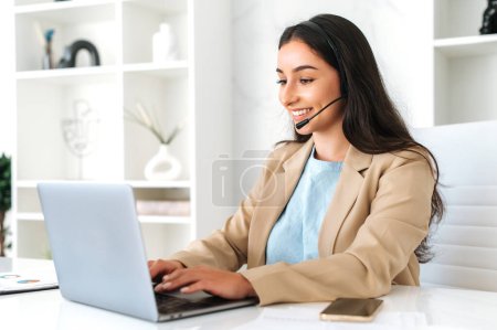 Téléchargez les photos : Femme amicale positive, opérateur de centre d'appels avec casque, travaillant sur la hotline de soutien dans un bureau moderne avec un ordinateur portable, regardant l'écran, souriant. Centre d'appels, service à la clientèle - en image libre de droit