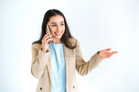 Téléchargez les photos : Femme brune indienne ou arabe positive en costume élégant, ayant une conversation de téléphone intelligent, geste à la main, debout sur un fond blanc isolé, regardant ailleurs, souriant joyeusement - en image libre de droit