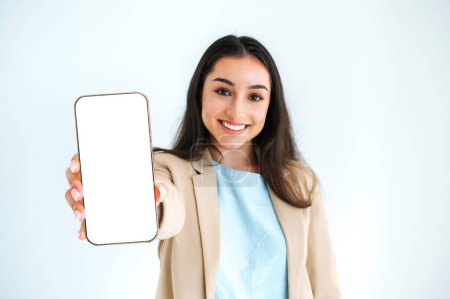 Mock-up-Bildschirm. Glückliche indische oder arabische Frau im eleganten Anzug, zeigt Smartphone mit leerem weißen Werbebildschirm, steht auf isoliertem weißem Hintergrund, blickt in die Kamera, lächelt