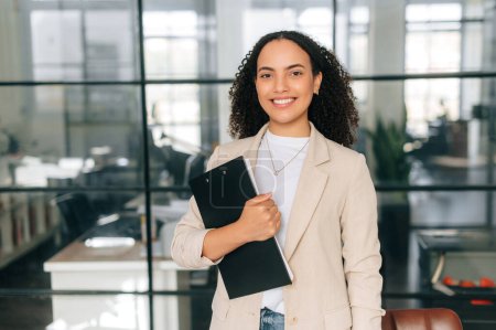 Motivated successful proud pretty, curly haired brazilian or hispanic business woman, corporate financial manager, teacher, in a suit, stand in modern office, holding documents, looks at camera, smile